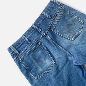 Puede incluir: Primer plano de la parte trasera de unos vaqueros azules. Los vaqueros tienen dos bolsillos traseros, trabillas y remaches de color cobre. El logotipo de Wrangler es visible en un peque&ntilde;o parche cerca del bolsillo trasero. El denim tiene un aspecto descolorido.