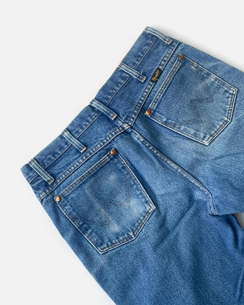 Puede incluir: Primer plano de la parte trasera de unos vaqueros azules. Los vaqueros tienen dos bolsillos traseros, trabillas y remaches de color cobre. El logotipo de Wrangler es visible en un peque&ntilde;o parche cerca del bolsillo trasero. El denim tiene un aspecto descolorido.