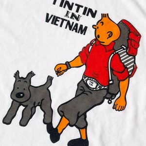 K&ouml;nnte beinhalten: Wei&szlig;es T-Shirt mit einer Cartoon-Illustration von Tim und Struppi, mit dem Text "TINTIN IN VIETNAM" in Schwarz. Tim wird mit Rucksack, rotem Hemd und grauer Hose dargestellt, wie er mit Struppi geht.