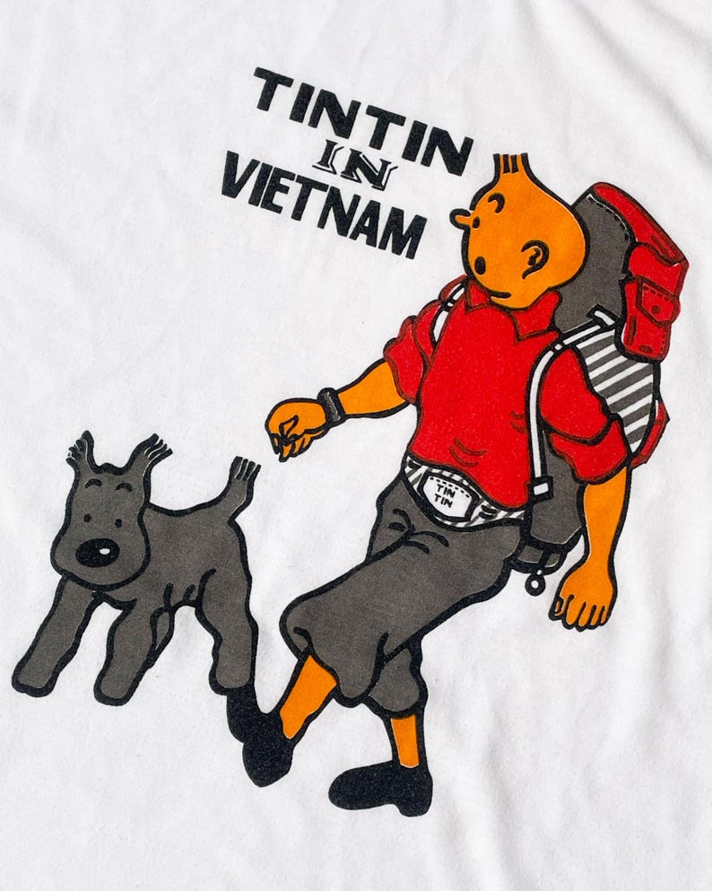 K&ouml;nnte beinhalten: Wei&szlig;es T-Shirt mit einer Cartoon-Illustration von Tim und Struppi, mit dem Text "TINTIN IN VIETNAM" in Schwarz. Tim wird mit Rucksack, rotem Hemd und grauer Hose dargestellt, wie er mit Struppi geht.