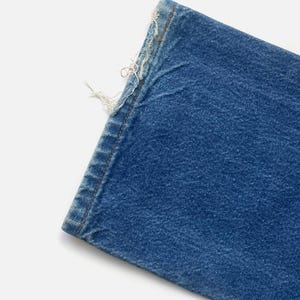 May include: Close-up of a section of blue denim jeans. The edge of the fabric is frayed, with visible threads and a worn appearance. The denim has a classic, medium-blue wash.