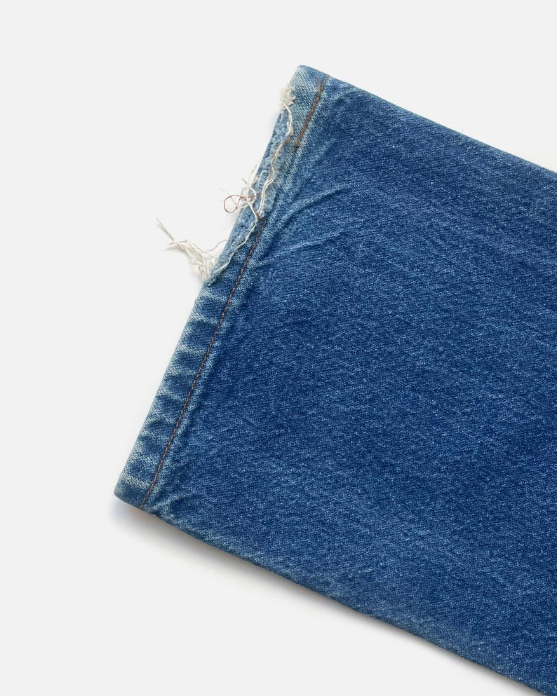May include: Close-up of a section of blue denim jeans. The edge of the fabric is frayed, with visible threads and a worn appearance. The denim has a classic, medium-blue wash.
