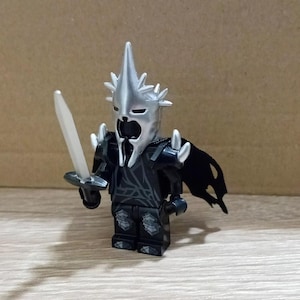 Puede incluir: Una minifigura Lego negra y plateada del personaje de El Señor de los Anillos, el Rey Brujo de Angmar, sosteniendo una espada plateada. La figura tiene una capa negra y un casco plateado con una corona puntiaguda.
