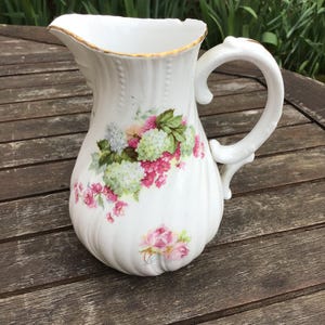 Peut inclure: Pichet en céramique blanche avec un bord doré et un motif floral. Le pichet a une anse et un corps arrondi. Les fleurs sont roses, blanches et vertes.