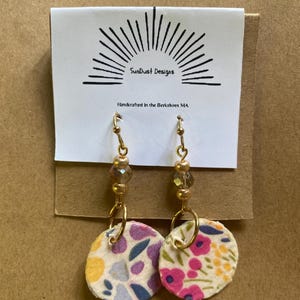 May include: A pair of dangle earrings with fabric circles featuring floral patterns in pink, purple, yellow, and blue. The earrings have gold-colored hoops and beaded accents. The card behind the earrings reads "SunDust Designs" and "Handcrafted in the Berkshires MA."