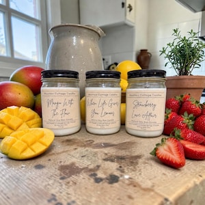 May include: Three glass jars of soy wax candles with black lids and labels that read "Mango With The Flow", "When Life Gives You Lemons", and "Strawberry Love Affair". The jars are surrounded by mangoes, lemons, and strawberries.