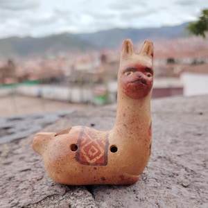 May include: A brown ceramic llama whistle with red and yellow geometric designs. The whistle has a hole on the side and is sitting on a stone surface.