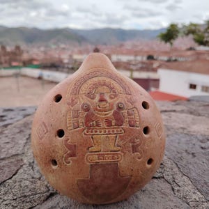 Puede incluir: Una ocarina de arcilla marrón con un diseño detallado de una figura con una corona y un patrón geométrico. La ocarina tiene seis agujeros para los dedos.