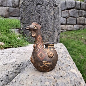 Puede incluir: Un jarrón de cerámica marrón con forma de pájaro, con un pequeño pico y asa. El cuerpo presenta patrones geométricos en rojo y marrón, con un diseño floral. El jarrón está sobre una superficie de piedra gris, con un fondo de muros de piedra y hierba verde.