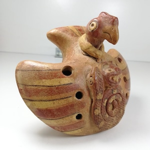 Puede incluir: Un silbato de terracota con forma de pájaro con una serpiente enroscada alrededor de su cuello. El pájaro tiene rayas rojas y amarillas en sus alas y una cabeza roja. La serpiente es marrón y tiene una superficie texturizada.