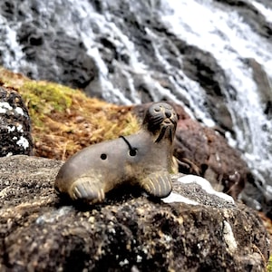 May include: A small, brown ceramic seal figurine with a black string tied around its neck. The figurine is sitting on a rock with a waterfall in the background.
