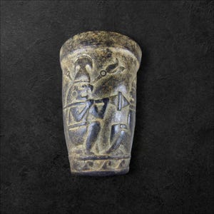 Puede incluir: Un jarrón de cerámica verde oscuro con un diseño tallado de dos figuras tocando instrumentos musicales. El jarrón tiene forma cilíndrica y un borde ligeramente acampanado.