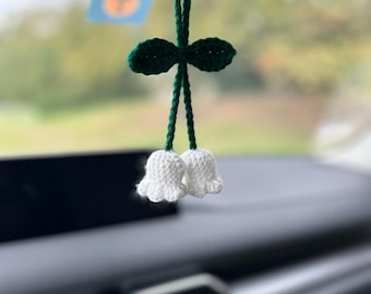 May include: A white crocheted flower with green leaves hanging from a rearview mirror in a car.