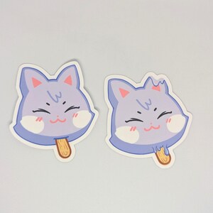 May include: Two stickers featuring a smiling cat face in the shape of a popsicle. The cat face is light purple with pink cheeks, a brown stick, and a white outline. The stickers are on a white background.