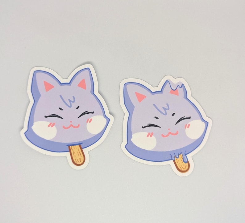 May include: Two stickers featuring a smiling cat face in the shape of a popsicle. The cat face is light purple with pink cheeks, a brown stick, and a white outline. The stickers are on a white background.