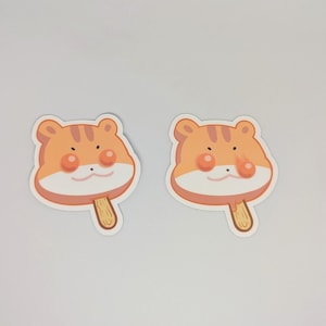 May include: Two stickers featuring a cartoon tiger face on a popsicle. The tiger face is orange with brown stripes, pink cheeks, and a small black nose. The popsicle stick is light brown.