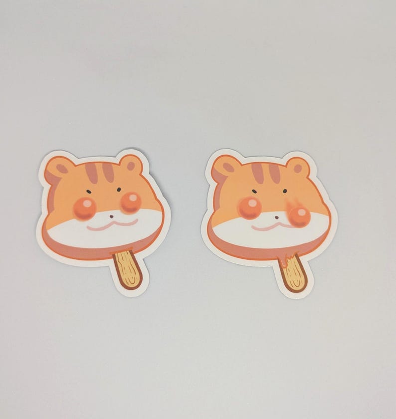 May include: Two stickers featuring a cartoon tiger face on a popsicle. The tiger face is orange with brown stripes, pink cheeks, and a small black nose. The popsicle stick is light brown.