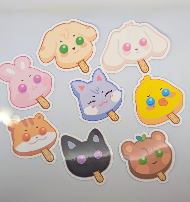May include: A collection of eight colorful stickers shaped like animal faces on popsicle sticks. The stickers feature a dog, bunny, cat, chick, hamster, and bear, each with a cute, cartoonish design and bright colors. The stickers are on a white surface.
