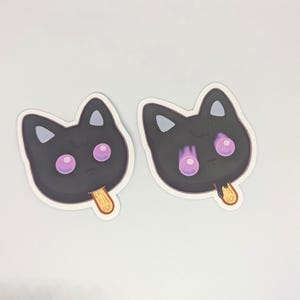 May include: Two stickers in the shape of a black cat face with pink eyes and a wooden popsicle stick. The cat's ears are triangular, and the eyes are large and round. The stickers are on a white background.