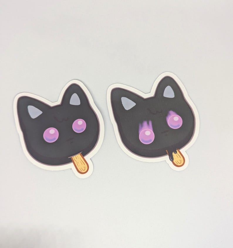 May include: Two stickers in the shape of a black cat face with pink eyes and a wooden popsicle stick. The cat's ears are triangular, and the eyes are large and round. The stickers are on a white background.