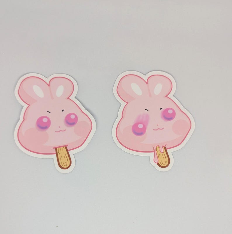 May include: Two pink bunny-shaped stickers resembling ice cream popsicles. Each sticker has a light pink face with rosy cheeks, dark eyes, and a small mouth. The popsicles have a wooden stick.