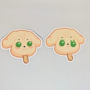 May include: Two stickers featuring a cartoon dog face with green eyes, a small black nose, and a light brown popsicle stick. The dog's face is light yellow with a light brown outline.