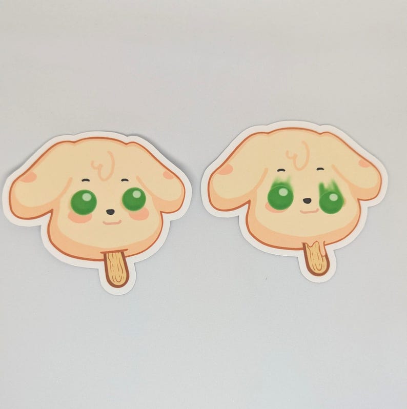 May include: Two stickers featuring a cartoon dog face with green eyes, a small black nose, and a light brown popsicle stick. The dog's face is light yellow with a light brown outline.