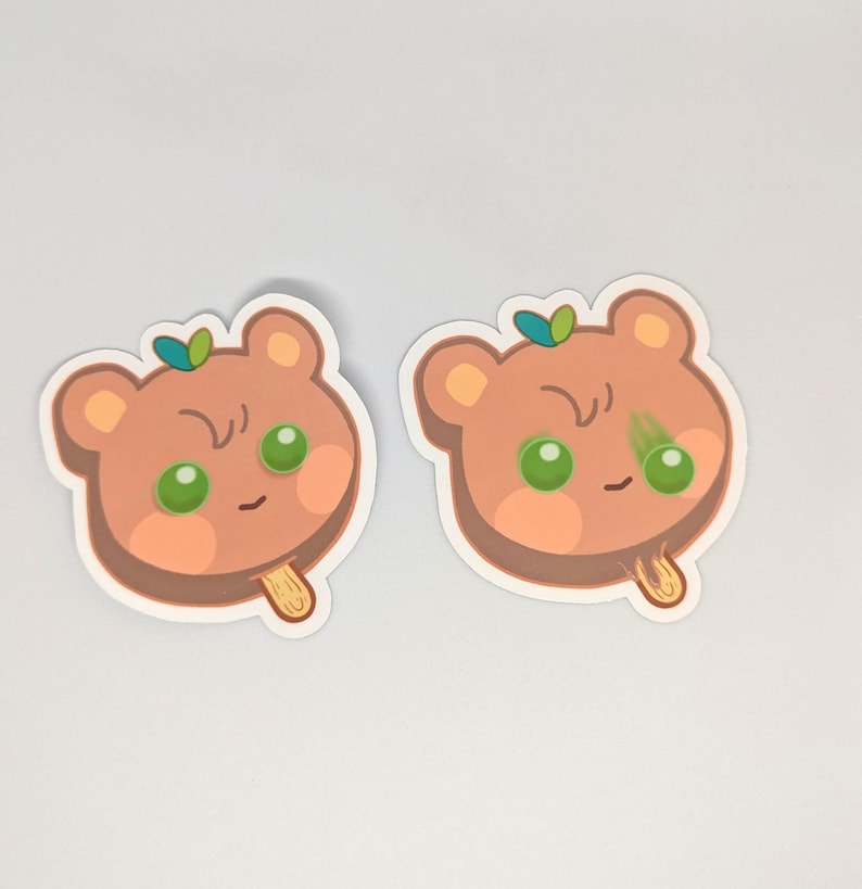 May include: Two stickers featuring a cartoon bear-shaped popsicle. The popsicles are light brown with green eyes and a small green leaf on top. The popsicle sticks are a light tan color.