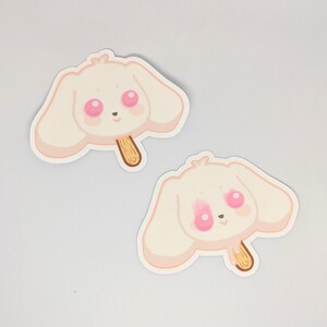 May include: Two stickers featuring a cartoon bunny head shaped like a popsicle. The bunny has large pink eyes and blush, with a light beige color and a wooden stick. The stickers are on a white background.