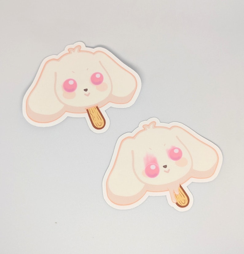 May include: Two stickers featuring a cartoon bunny head shaped like a popsicle. The bunny has large pink eyes and blush, with a light beige color and a wooden stick. The stickers are on a white background.
