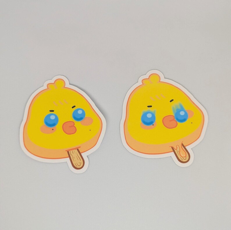 May include: Two stickers featuring a yellow chick design shaped like a popsicle. The chick has blue eyes, an orange beak, and a light orange outline. One chick has a single tear. The stickers have a wooden popsicle stick.