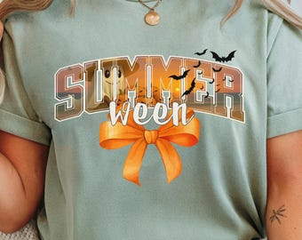 May include: Sage green t-shirt with the text "Summerween" in a brown and orange gradient font, featuring a pumpkin and bats. An orange bow is below the text. The shirt is worn by a person.
