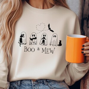 Könnte beinhalten: Ein sandfarbenes Sweatshirt mit einem Halloween-Motiv. Die Grafik zeigt Cartoon-Geister, eine Fledermaus und den Text "Boo & Mew". Eine orangefarbene Tasse wird gehalten. Ein Kürbis befindet sich im Hintergrund.