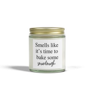 May include: A clear glass jar candle with a gold lid. The candle has a white label with the text "Smells like it's time to bake some sourdough" in black script. The candle is on a white background.