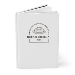 Op de afbeelding: Een wit dagboek met een zwart-witte illustratie van een brood op een bord. De tekst "BREAD JOURNAL 2025" staat op de omslag.
