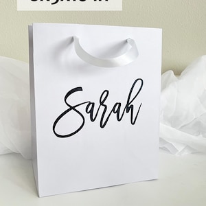 May include: White gift bag with the name "Sarah" in black script. The bag measures 8x5x10 inches and has a white ribbon handle. The bag is made of a sturdy material and is suitable for gifts.
