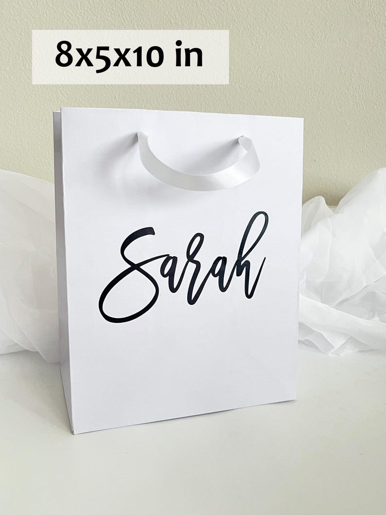 May include: White gift bag with the name "Sarah" in black script. The bag measures 8x5x10 inches and has a white ribbon handle. The bag is made of a sturdy material and is suitable for gifts.