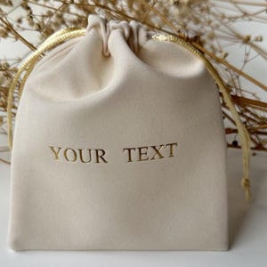 May include: A beige velvet drawstring bag with gold text that reads "YOUR TEXT". The bag has a gold drawstring closure and is set against a white background with dried floral accents. Ideal for small gifts.