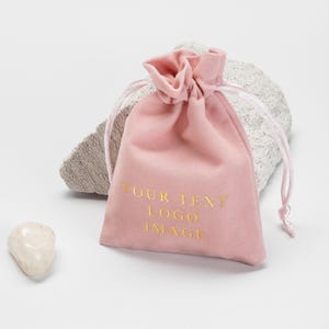May include: A soft pink velvet drawstring bag with the text "YOUR TEXT LOGO IMAGE" in gold. The bag is partially closed with a pink drawstring. A small white stone and a gray rock are in the background.