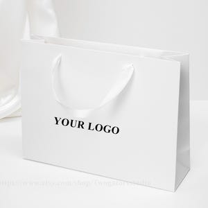 May include: A white rectangular shopping bag with a satin ribbon handle. The words "YOUR LOGO" are printed in black on the front of the bag. The bag is sitting on a white surface.