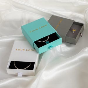 May include: Three jewelry boxes in white, teal, and gray, each with a drawer. The teal box contains a silver necklace with a blue heart pendant. The white box holds a silver bracelet. The gray box has a yellow gemstone ring. The text "YOUR LOGO" is printed on the boxes.