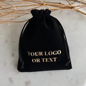 May include: Black velvet drawstring bag with gold text that reads "YOUR LOGO OR TEXT". The bag is closed with a black drawstring. The background is a white marble surface with dried floral accents.