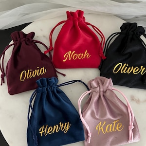 May include: Five velvet drawstring bags in shades of burgundy, red, black, navy blue, and mauve. Each bag features a gold embroidered name: Olivia, Noah, Oliver, Henry, and Kate. The bags are displayed on a white marble surface.