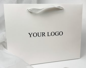 May include: A white gift bag with a satin handle and the text "YOUR LOGO" printed in black. The bag is rectangular and sits on a white surface with white fabric draped around it.
