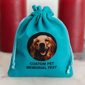 May include: A teal drawstring bag with a circular image of a golden retriever dog. The bag has the text "CUSTOM PET MEMORIAL TEXT" printed below the image. Two red candles are in the background.