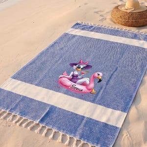 May include: A rectangular blue and white beach towel with a geometric diamond pattern and fringe on the ends. The center features a graphic of a cartoon duck wearing a purple hat and dress, sitting in a pink flamingo float with the name Stella.