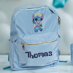 May include: A light blue backpack and matching pencil case featuring a cartoon character design. The backpack and pencil case have gold zippers and the name "Thomas" printed on them. The background includes decorative stars and clouds.
