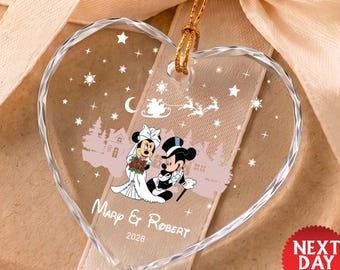 May include: A clear heart-shaped glass ornament featuring a printed graphic of two cartoon mice dressed in wedding attire, a silhouette of a sleigh with reindeer, stars, and snowflakes. The text reads Mary & Robert 2026 in white lettering.