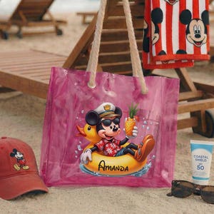 May include: A red baseball cap with a Mickey Mouse graphic, a pink tote bag with a Mickey Mouse design, a red and white striped towel, sunglasses, and sunscreen on a sandy beach. The tote bag features Mickey Mouse in a duck floatie.