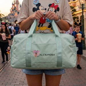 Może przedstawiać: Jasnozielona torba podr&oacute;żna z uchwytami i paskiem na ramię. Torba ma kieszeń z grafiką Minnie Mouse i nadrukowanym pod spodem imieniem "ALEXANDRA". Do torby przymocowany jest brelok do kluczy z Myszka Miki. Osoba ma na sobie koszulkę Disneya.